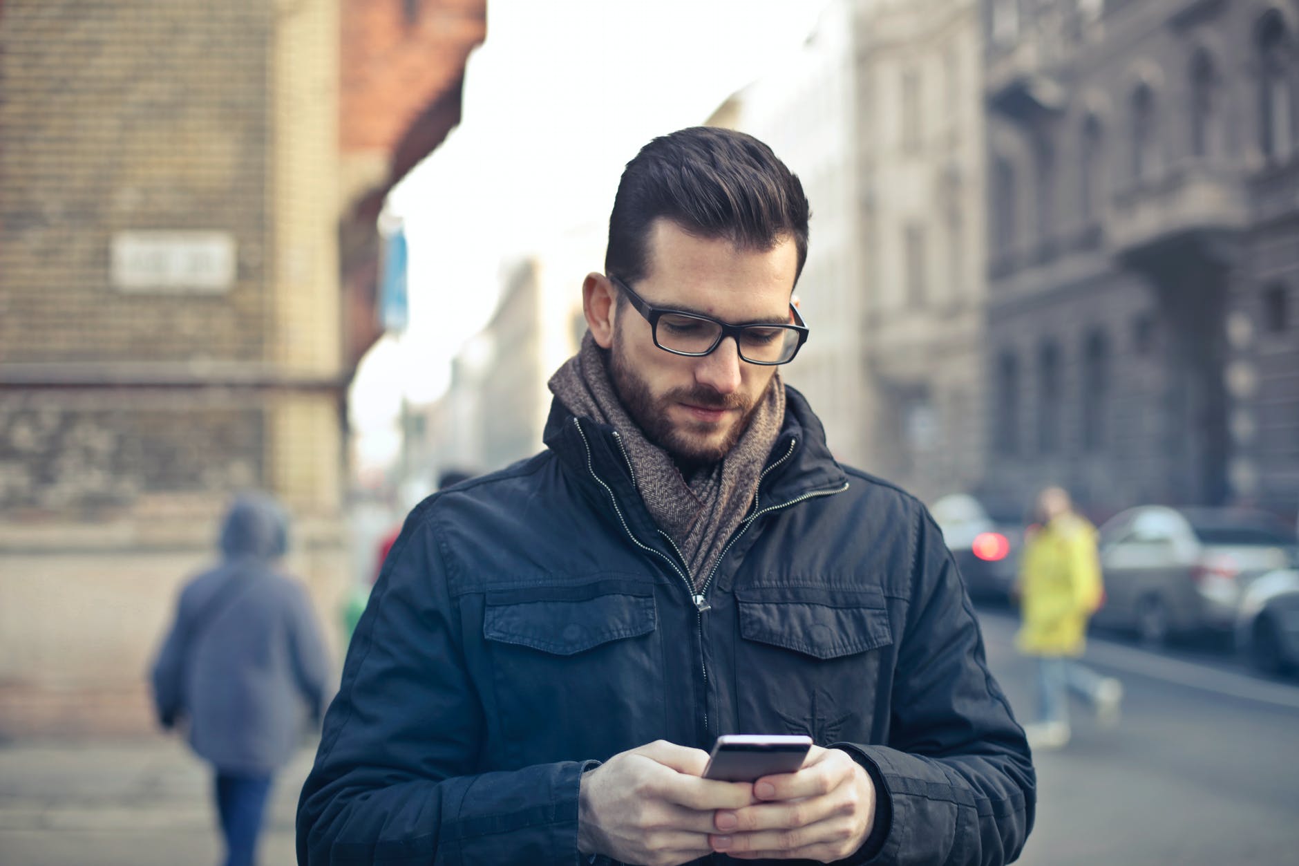 man wearing black zip jacket holding smartphone surrounded by grey concrete buildings